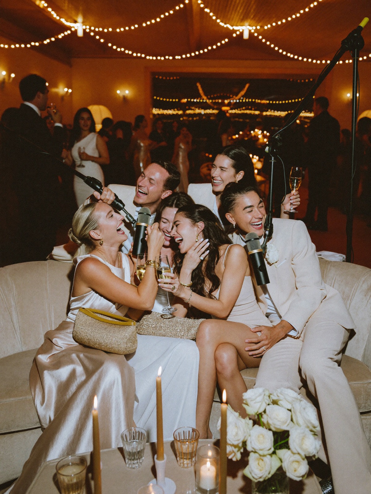 Wedding guests laughing together into podcast microphones on a velvet sofa, the reception glowing behind them